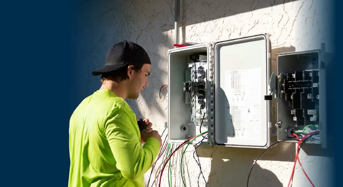 Electrician in a neon green shirt and black cap working on an open outdoor electrical panel.