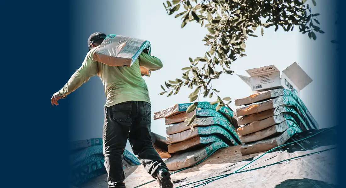Man carrying a roofing material bag on his shoulder while walking on a roof with stacked bundles nearby.