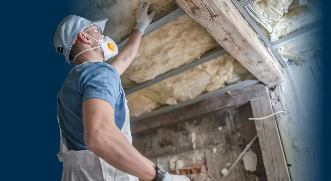 Worker in mask and gloves installing insulation in a ceiling under wooden beams.