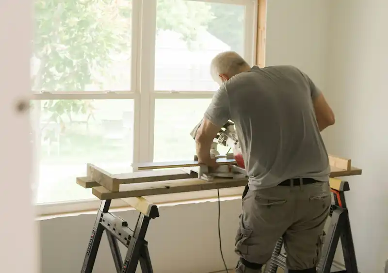 Man using a miter saw to cut wood indoors near a window on a workbench with sawhorse supports.