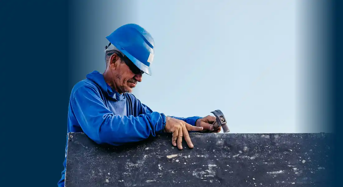 Worker in blue uniform and helmet using hammer on a dark surface against sky background.