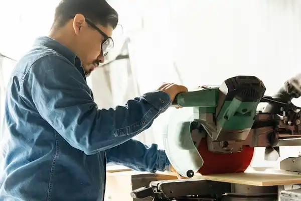 Man wearing glasses and denim shirt operating a green power saw cutting wood in workshop.