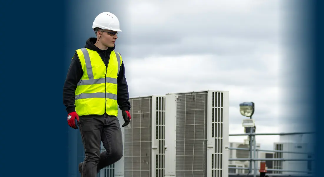 Construction worker in safety vest and helmet inspecting rooftop HVAC units under cloudy sky.