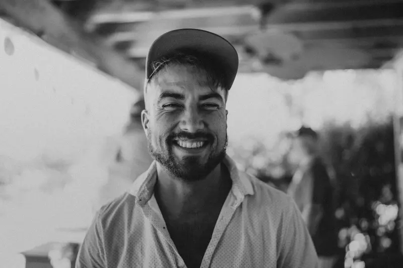 Smiling man wearing a cap and buttoned shirt in a blurred outdoor setting.