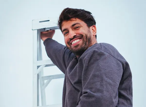 Smiling man in a gray sweater climbing a white ladder against a light background.