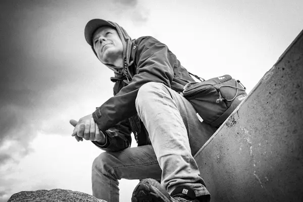Black and white photo of a man sitting outdoors wearing a cap and jacket, looking up thoughtfully.