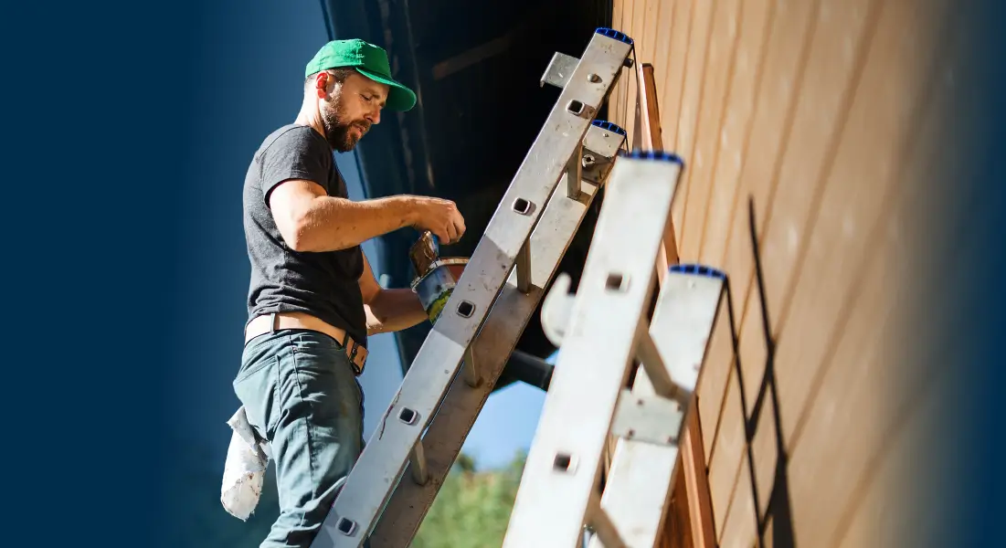 A worker on a ladder holding a paintbrush and paint near a wooden wall.