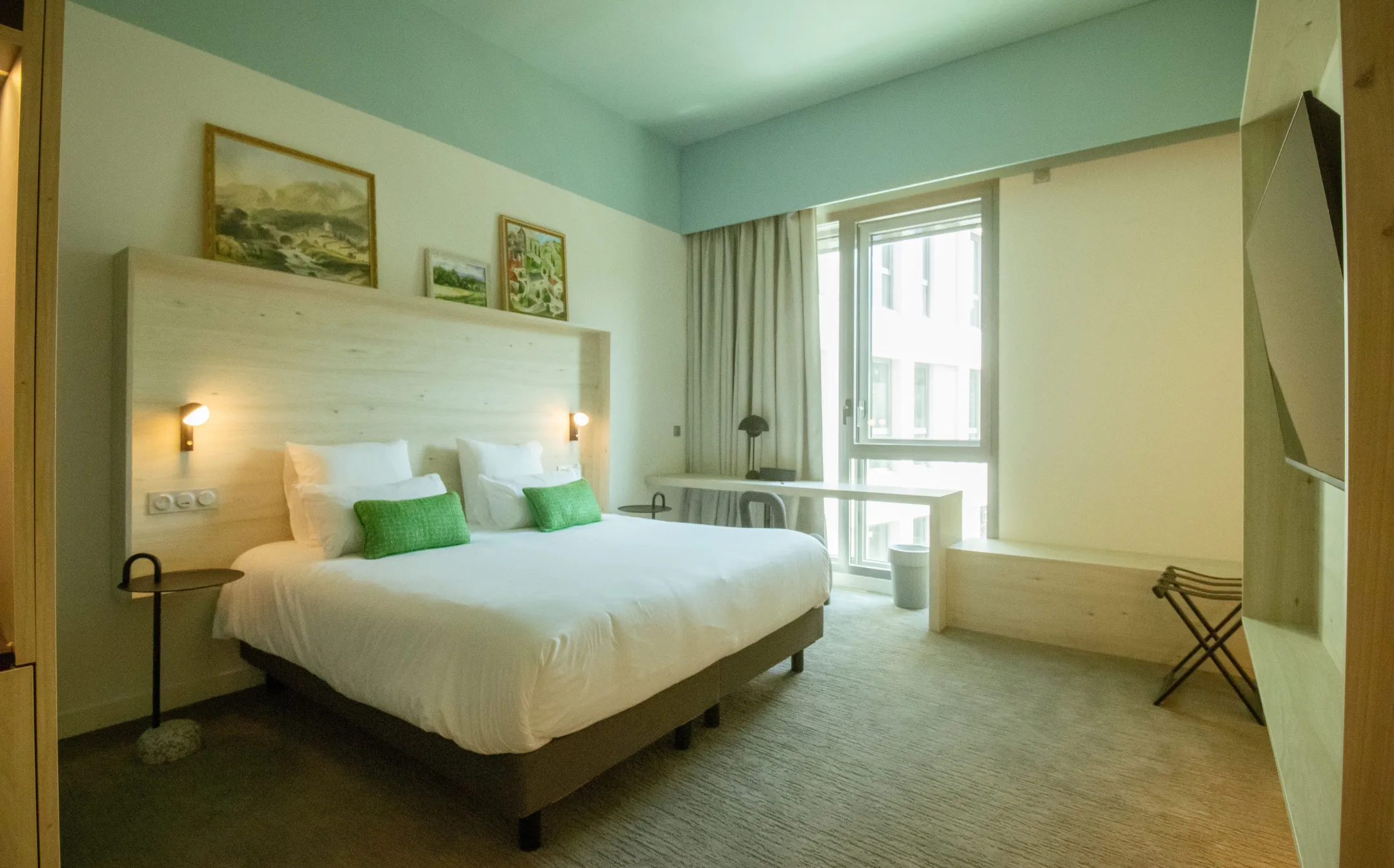 Bright bedroom with large double bed, green cushions, desk near a large window.