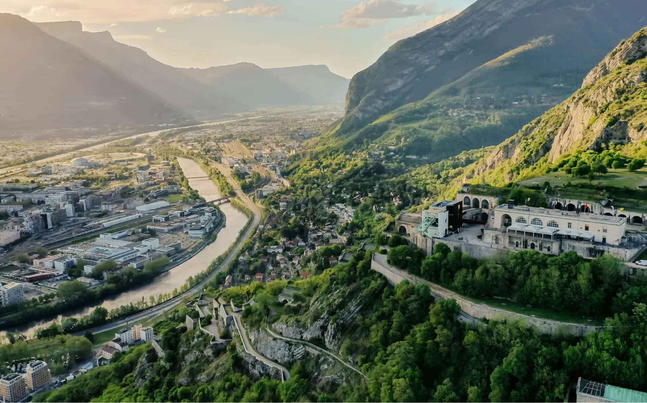 Vue aérienne de Grenoble avec une rivière, des montagnes et une forteresse sur une colline verdoyante.