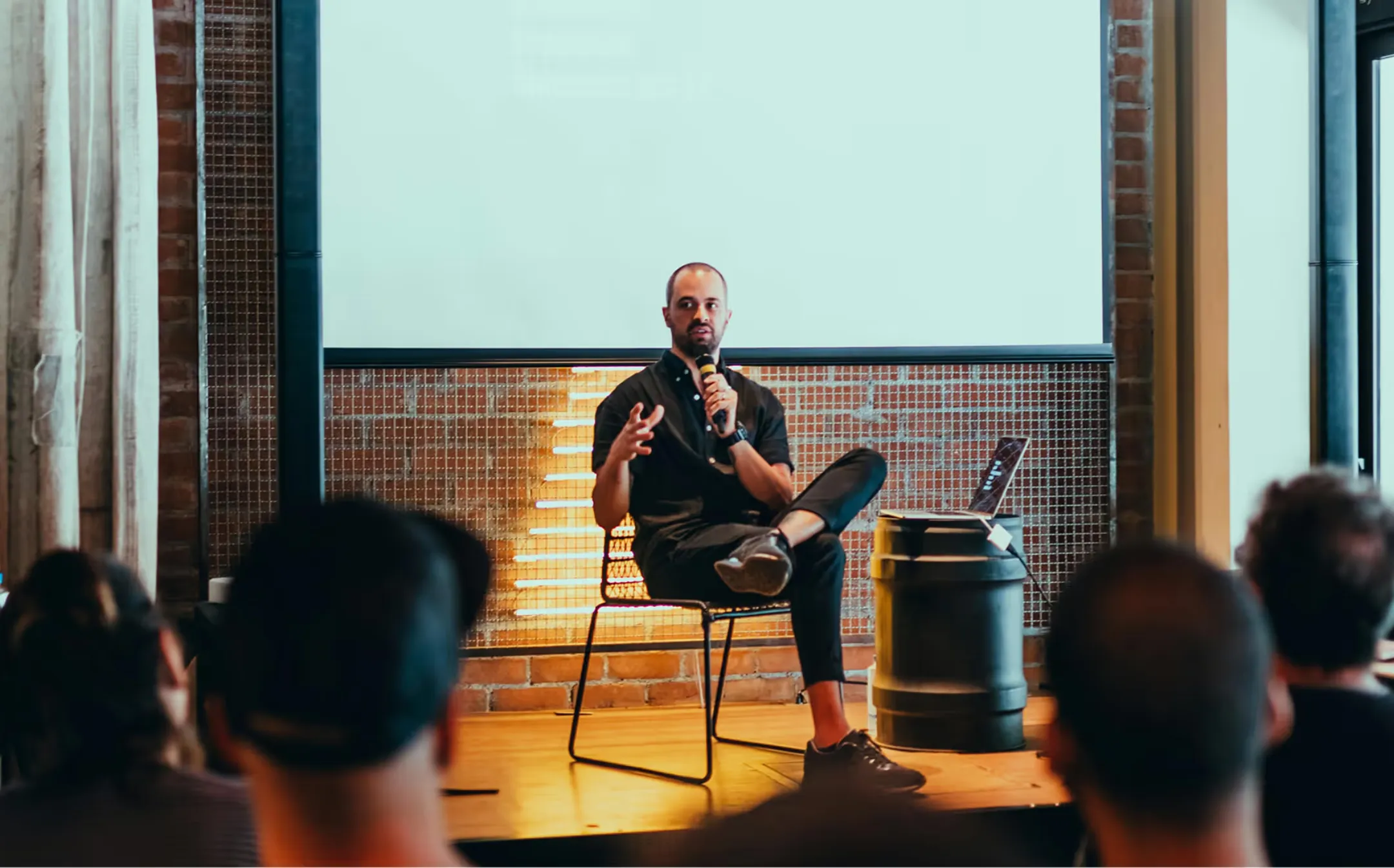 Man sitting with microphone speaking in front of an audience in a room with screen and laptop.