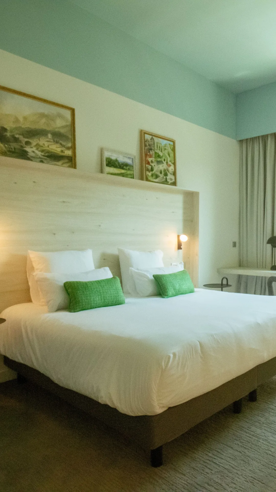 Hotel room with double bed, white pillows and green cushions, minimalist decor with paintings.