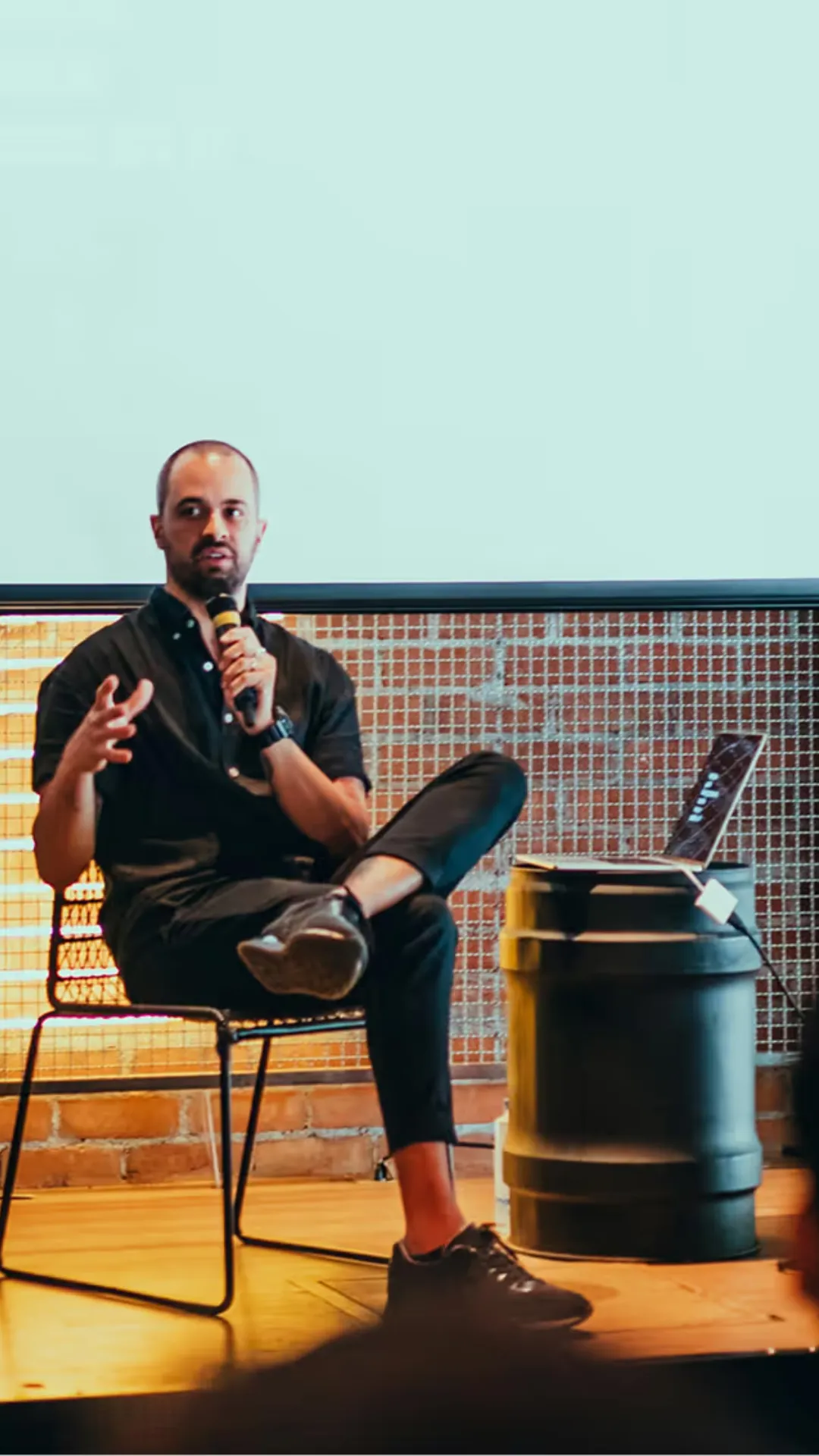 Man talking to the microphone sitting on a chair, with a laptop on a barrel next to it.
