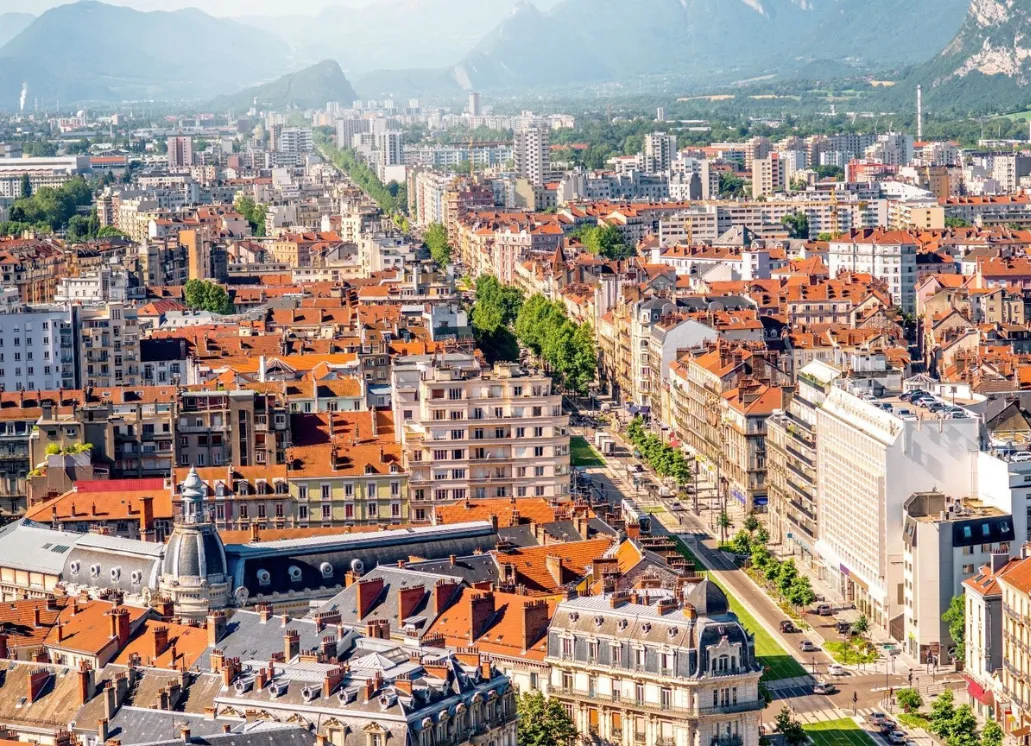 Aerial view of a city with red roofs, tree-lined streets and mountains in the background.