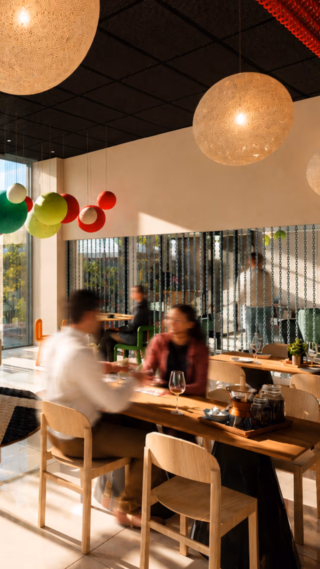 Deux personnes floues discutent à une table en bois dans un café lumineux avec des lampes et des décorations.