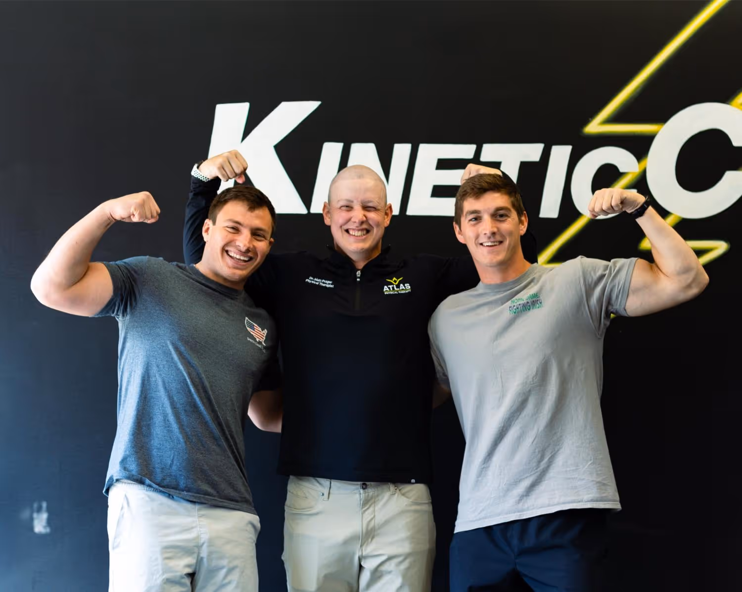 Three men smiling and flexing their arm muscles standing in front of a wall with 'KINETIC' text.
