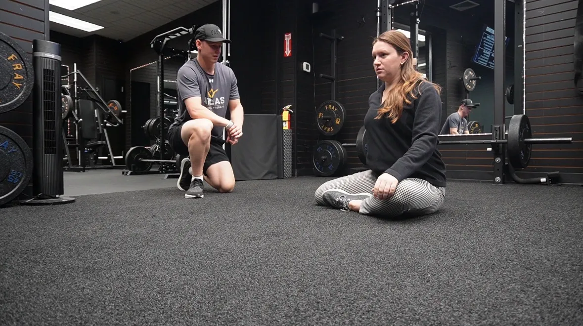 Man kneeling talking to woman sitting cross-legged on gym floor with weightlifting equipment behind.