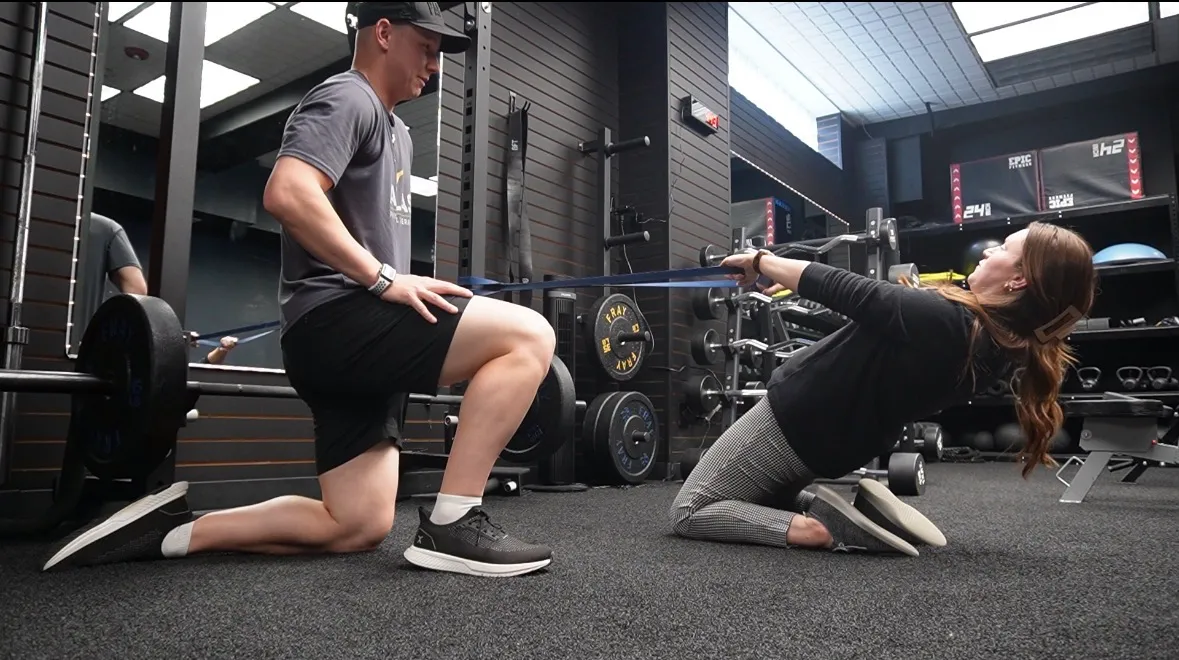 Man assisting woman with resistance band exercise in gym surrounded by weights and equipment.