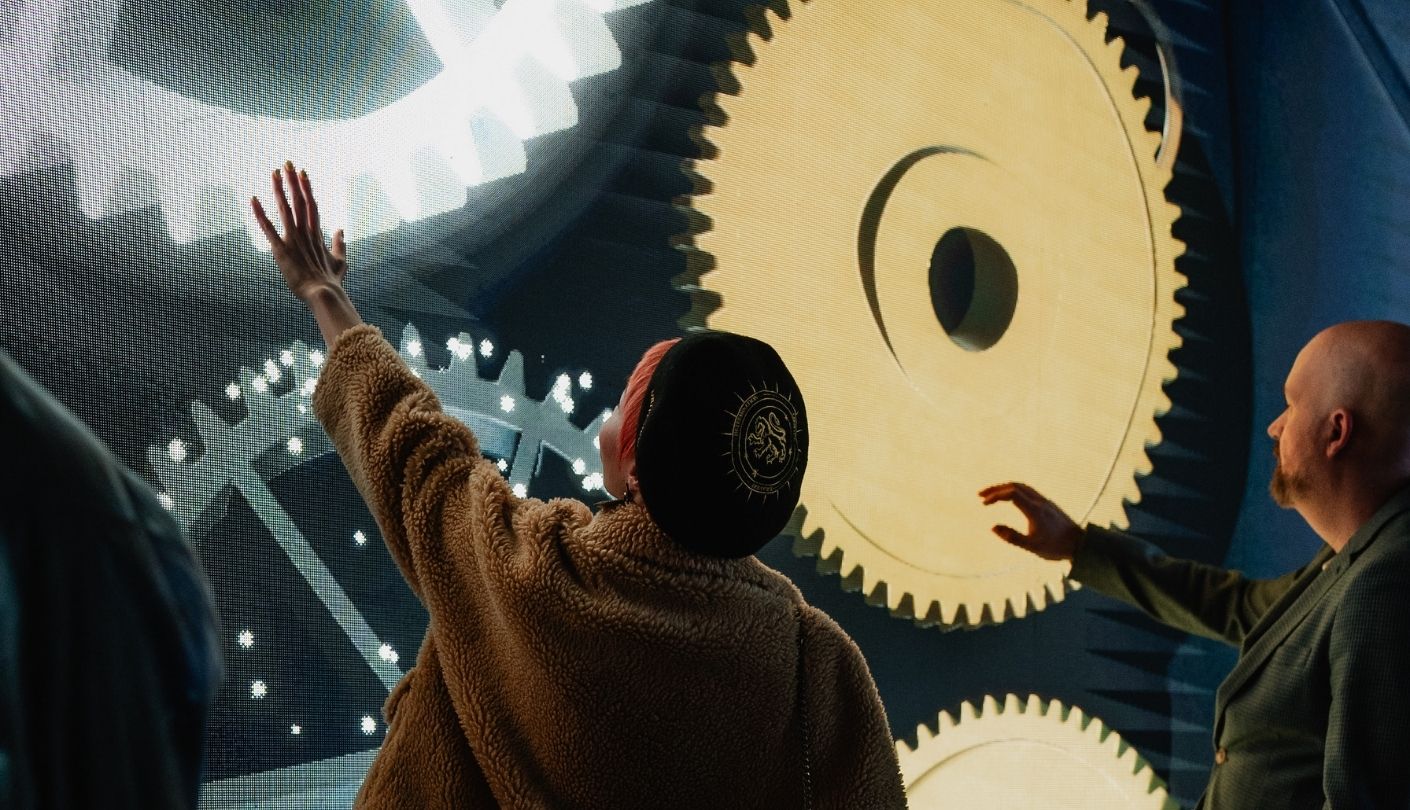 Two visitors reach toward glowing gears on a curved digital screen