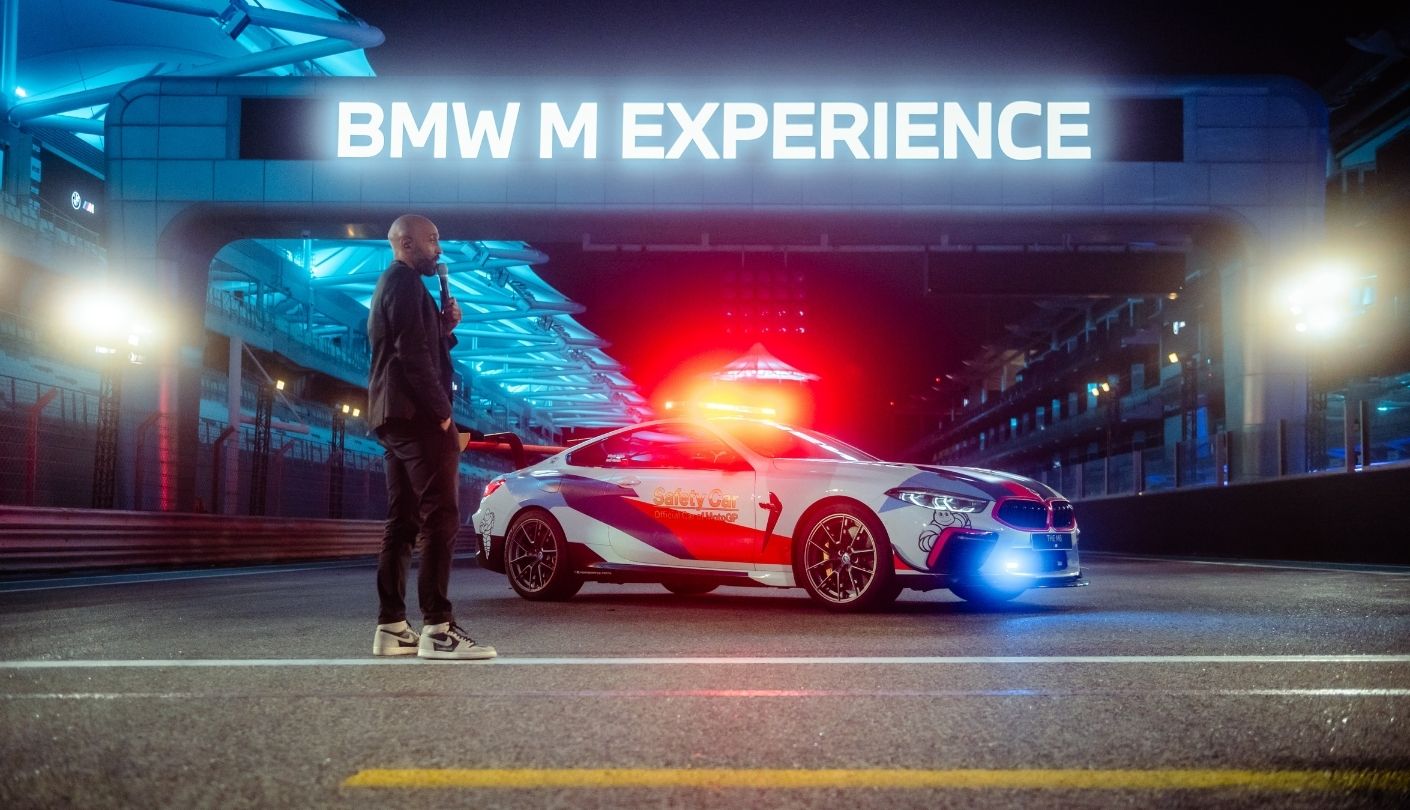 Presenter beside a BMW M safety car under a neon experience sign