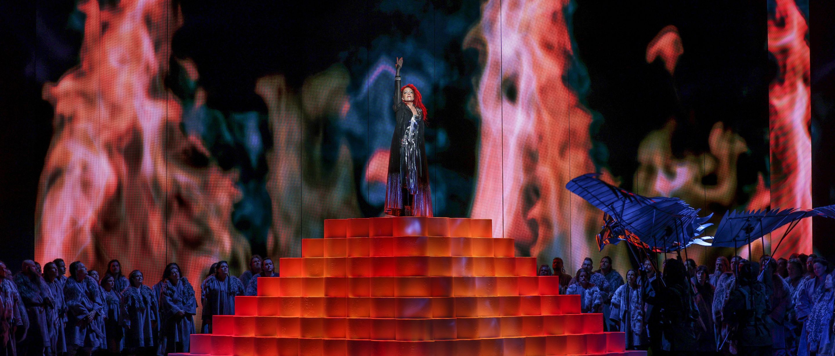 Singer stands on a red pyramid before a fiery opera stage backdrop