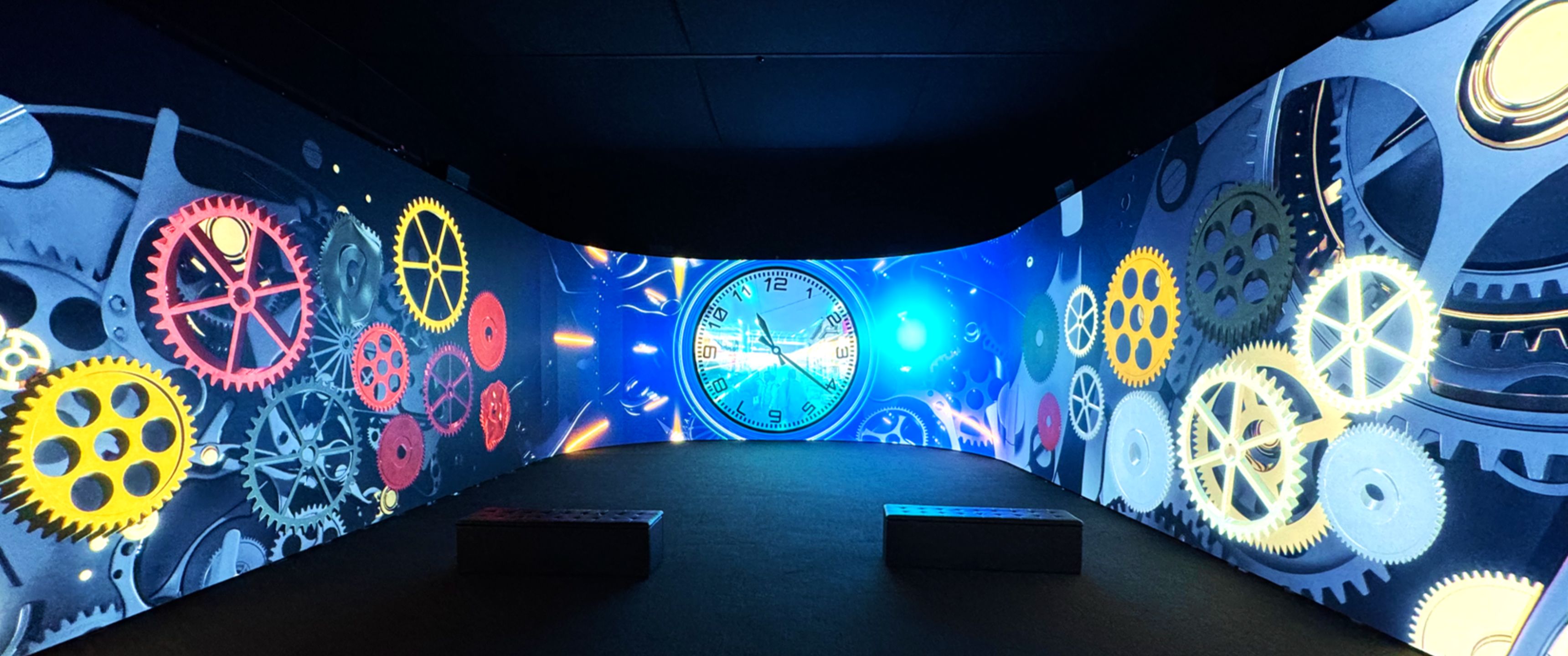 Curved screens show colorful gears and a large clock in dark room