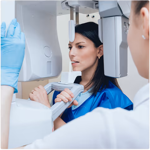 Woman wearing protective lead apron undergoing dental X-ray with a technician operating the machine.