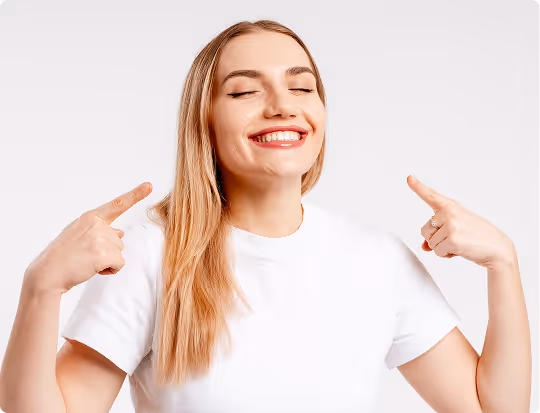 Smiling woman with long blonde hair pointing fingers at her cheeks while wearing a white t-shirt.