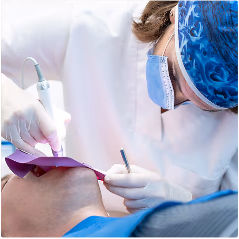 Dentist wearing a surgical mask and cap performing a dental procedure on a patient’s mouth.