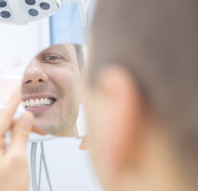 Man smiling and checking his teeth in a dental mirror at the dentist's office.