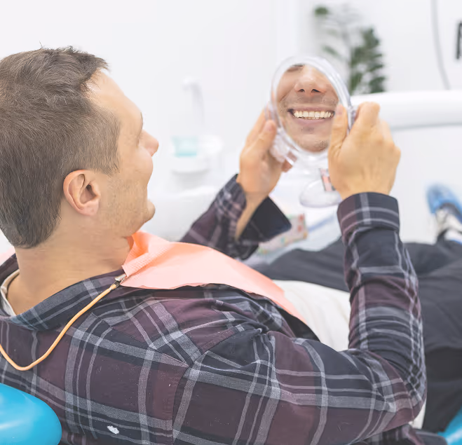 Man sitting in dental chair smiling and looking at his teeth in a handheld mirror.