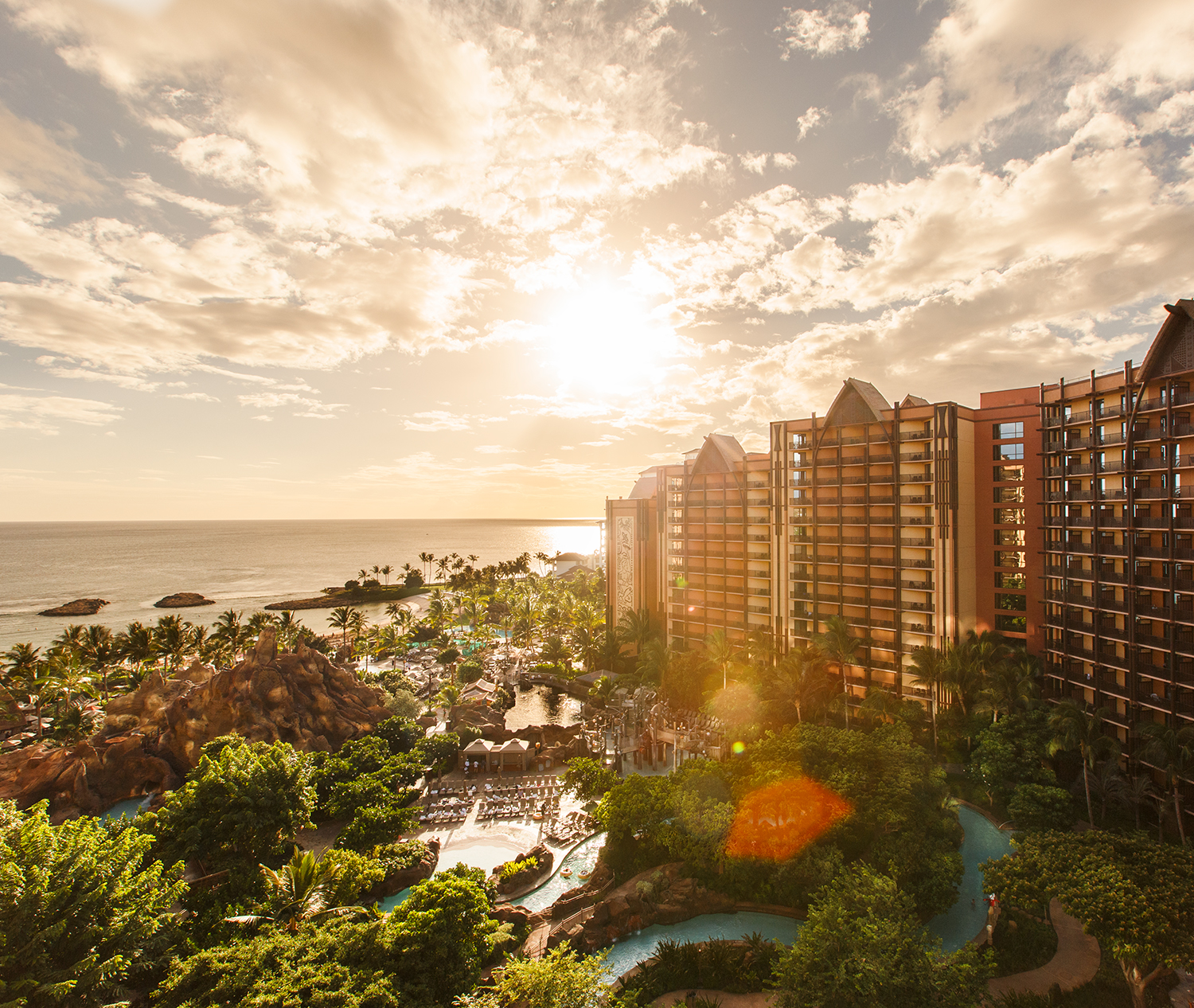 Sunset over a resort with pools, lush greenery, and ocean view under partly cloudy sky.
