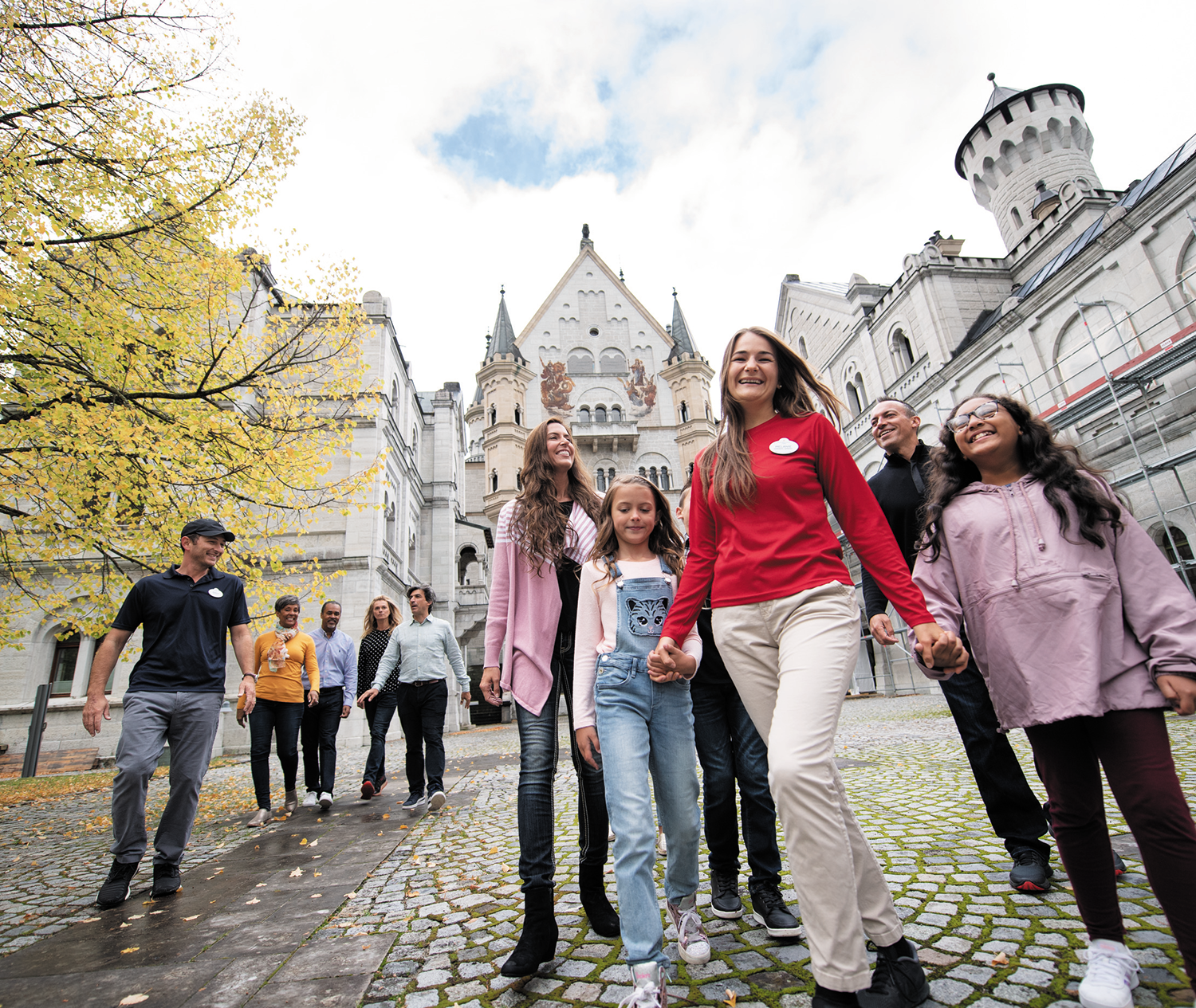 Group of people walking happily on a cobblestone path near a castle with autumn trees on the side.