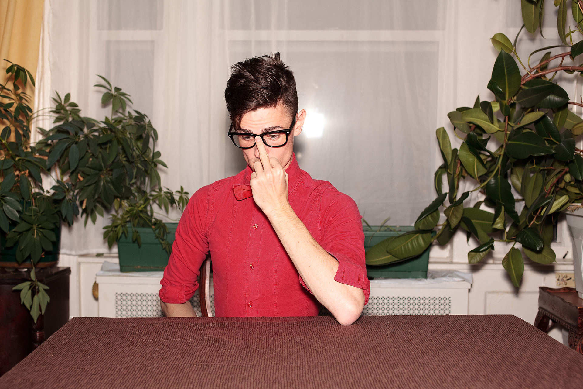 Person in red shirt and glasses adjusting their glasses while sitting at a table with plants behind them.