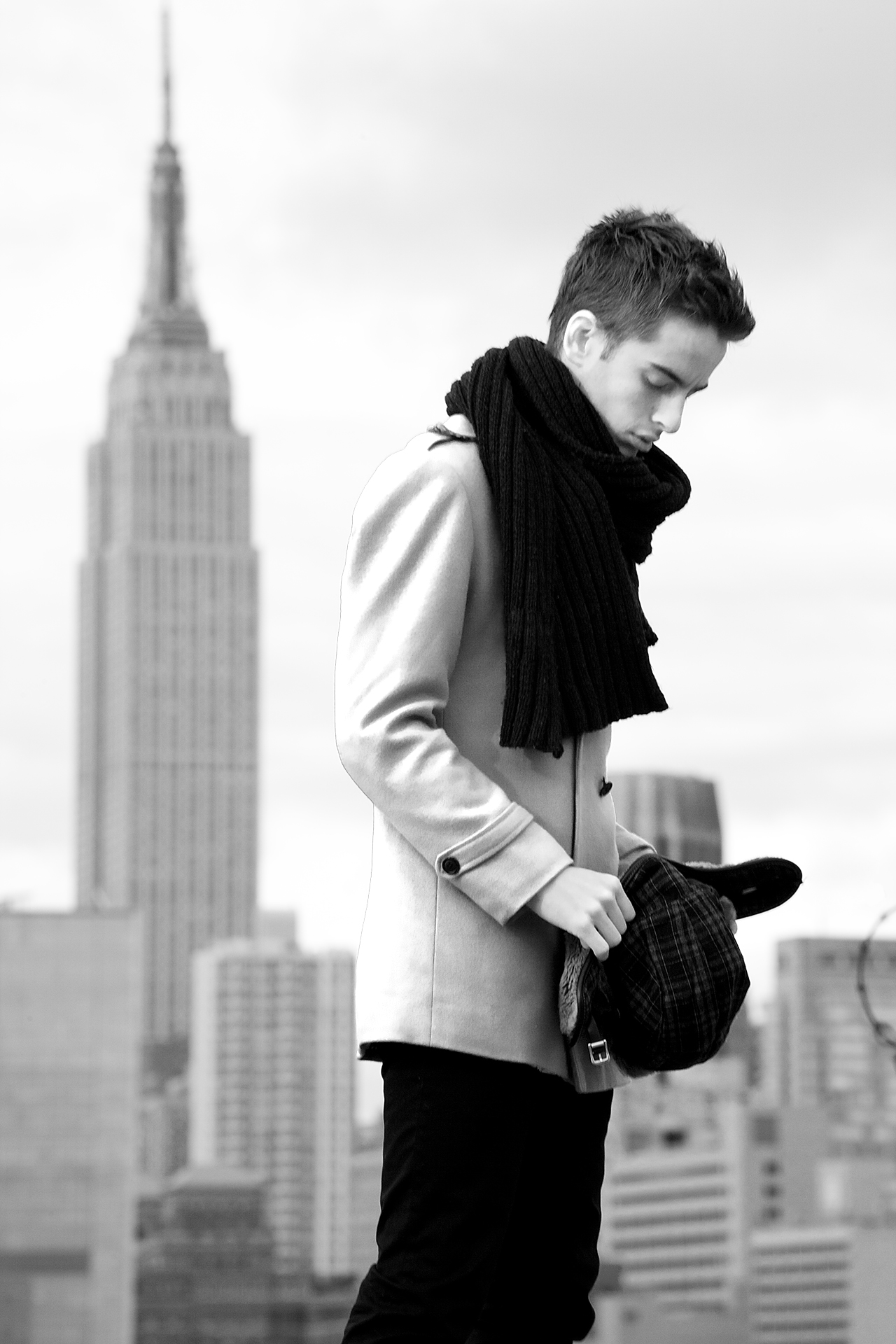 Young man in a coat and scarf holding a hat, with a blurred city skyline behind him.