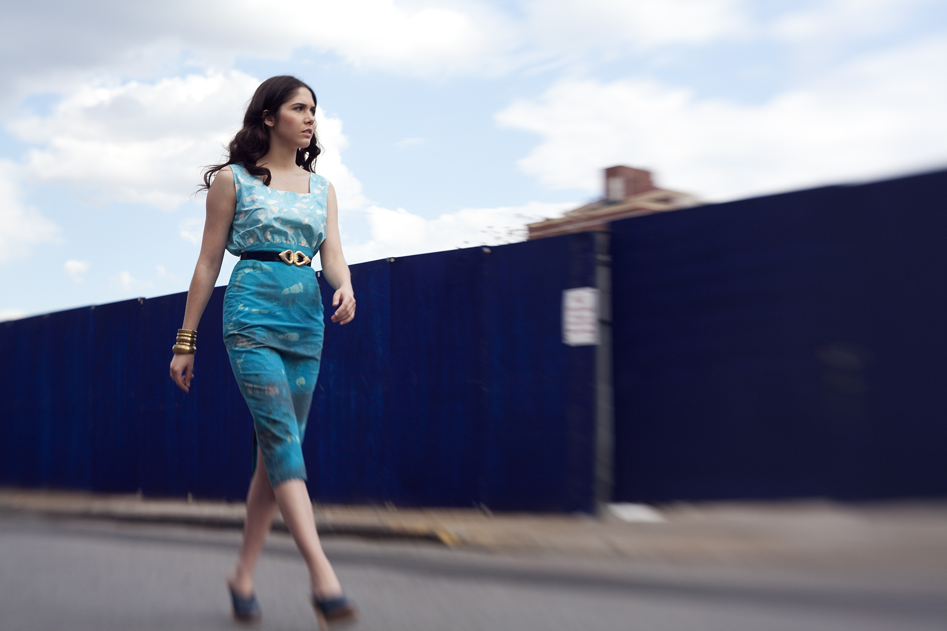 Woman walking on street wearing a fitted turquoise dress with gold belt and bracelets.