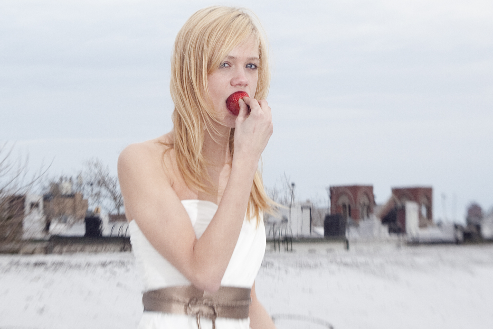 Blonde woman in white dress biting a strawberry on a rooftop with a cloudy sky background.