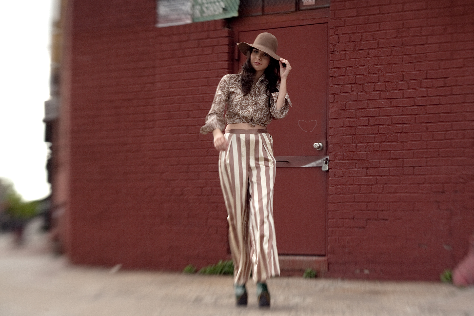 Woman in patterned blouse, striped pants, and wide-brimmed hat standing in front of a red brick wall.