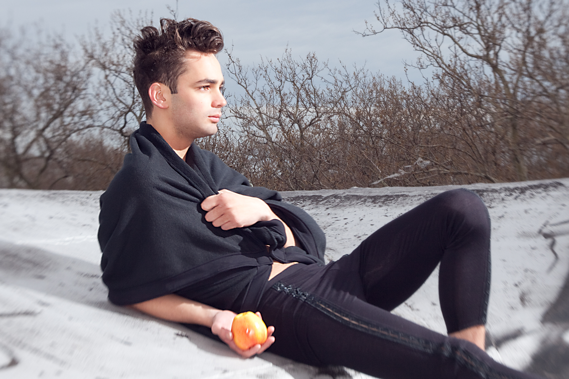 Young man in black wrapped in a blanket holding an apple outdoors with bare trees behind him.