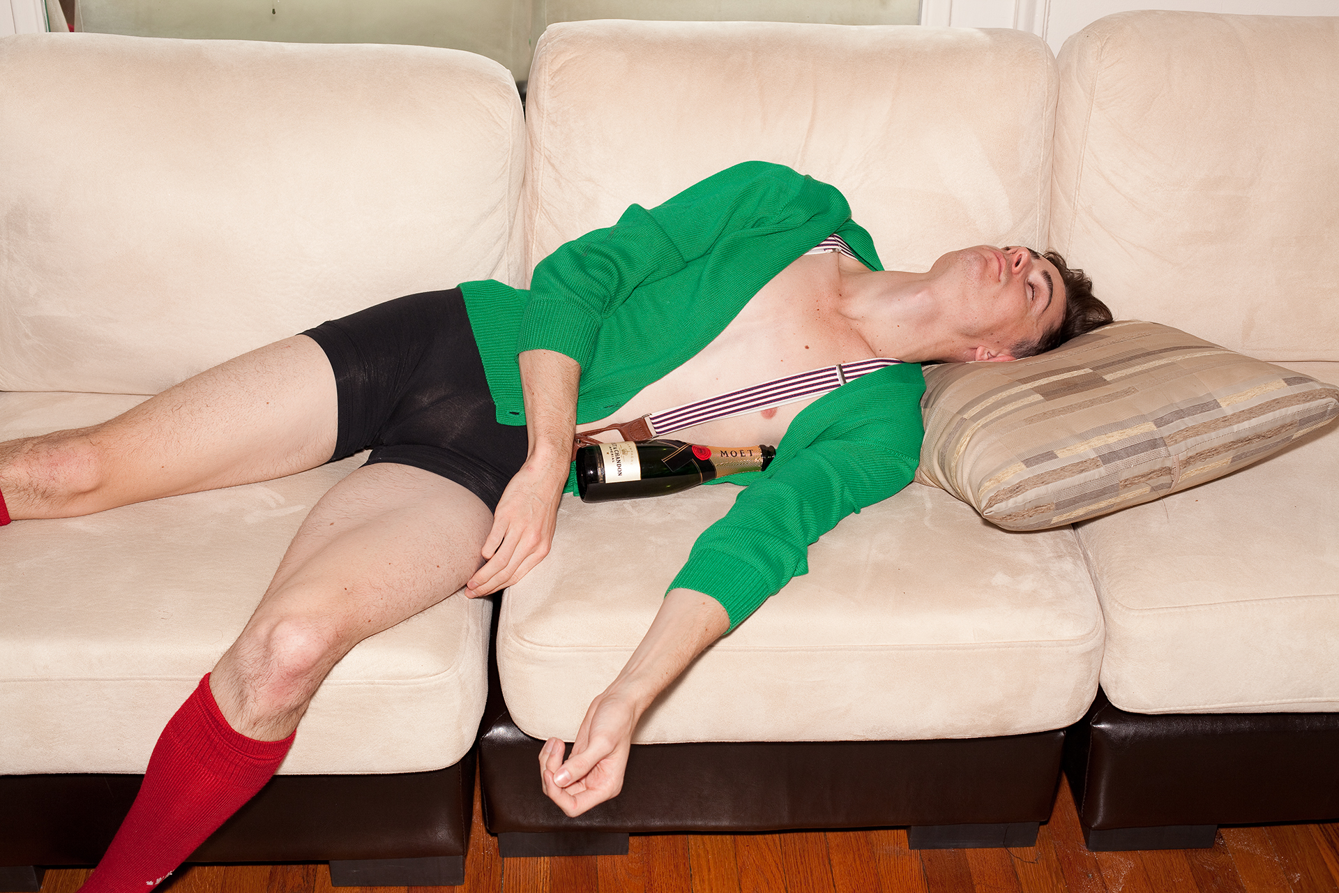 Man lies on beige couch with bottle of champagne under arm, wearing green sweater, black shorts, red socks.