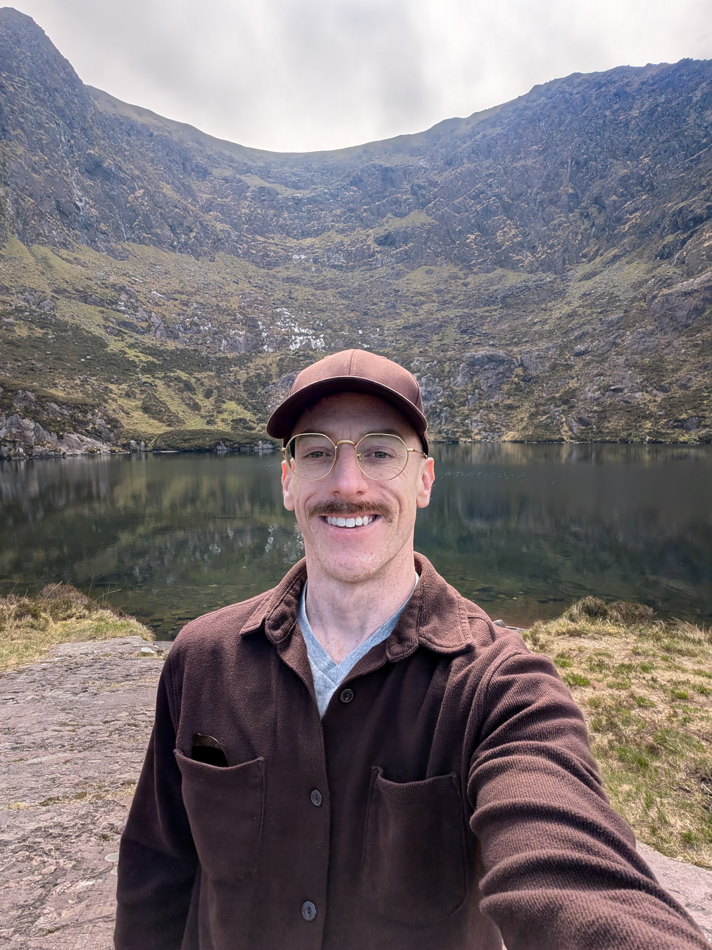 A man in a brown jacket and glasses is taking a picture of himself in front of a mountain.