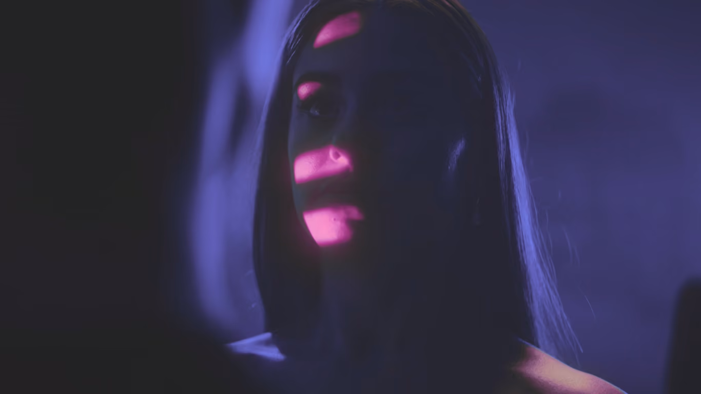 A woman's face partly illuminated by purple and pink light stripes in a dark, moody setting.