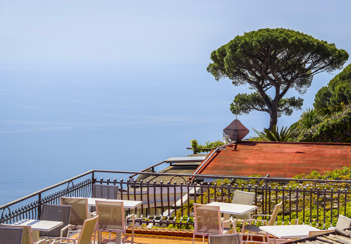 Outdoor terrace with empty tables and chairs overlooking calm sea and large tree on a sunny day.