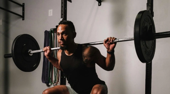 A man squats with a barbell in a gym, conveying strength and focus. He wears a black tank top, surrounded by gym ropes and equipment, emitting determination.
