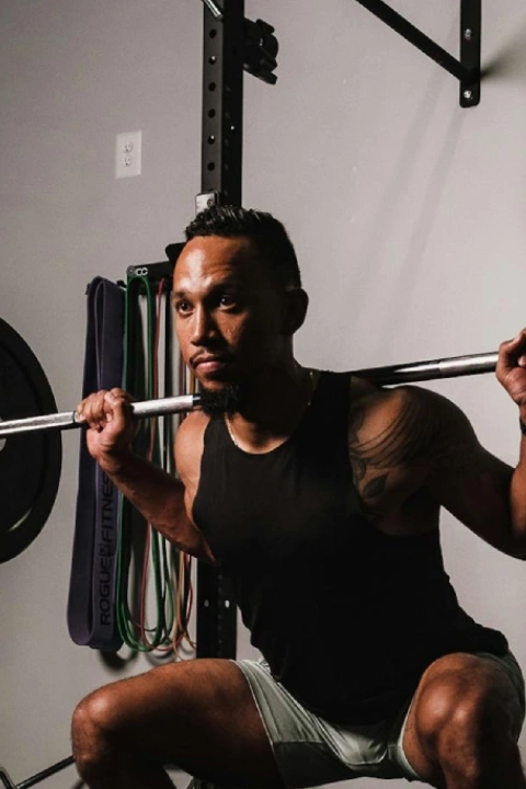 A man squats with a barbell in a gym, conveying strength and focus. He wears a black tank top, surrounded by gym ropes and equipment, emitting determination.