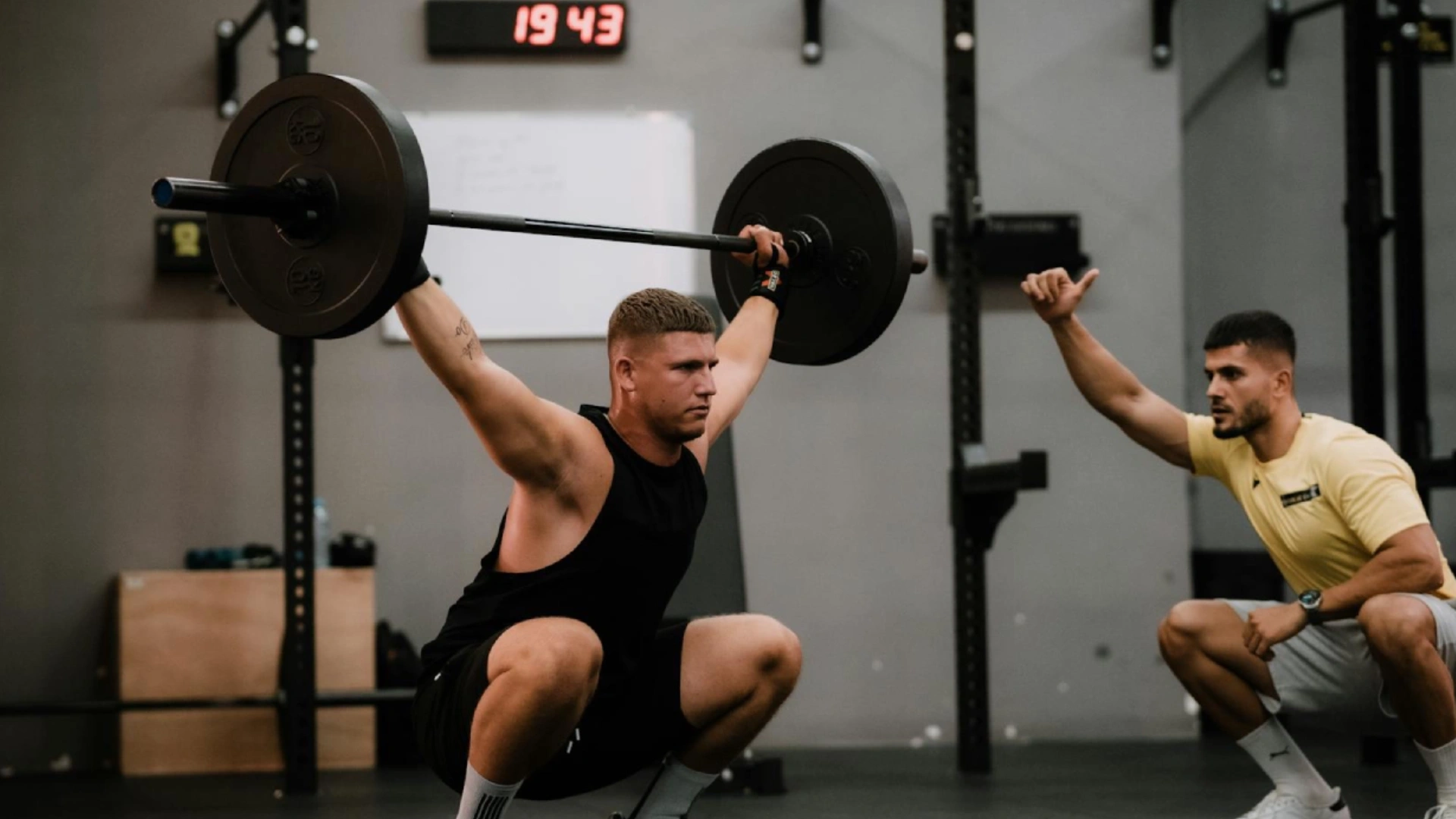 A man in gym attire performs a barbell squat in a gym, while a coach in a yellow shirt kneels beside him, giving a thumbs-up. A timer on the wall reads 19:43.
