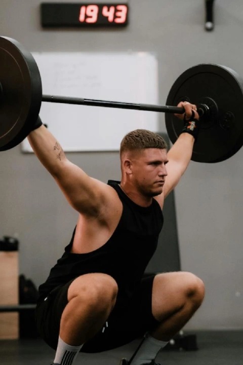 A man in gym attire performs a barbell squat in a gym, while a coach in a yellow shirt kneels beside him, giving a thumbs-up. A timer on the wall reads 19:43.