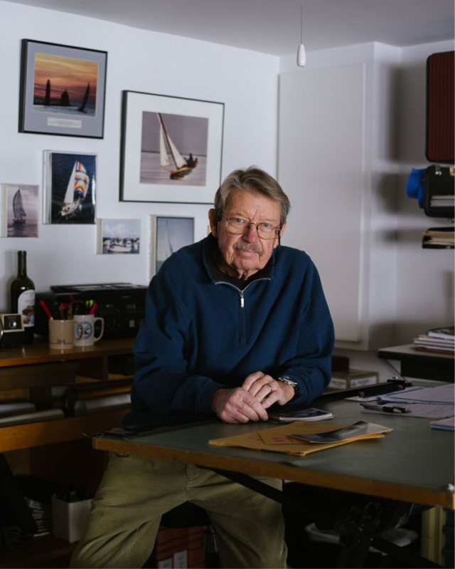 Older man wearing glasses and a navy blue pullover sitting at a desk with papers and photos of sailboats on the wall behind him.