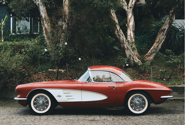 Side view of a classic red and white vintage convertible car parked on a street with trees and bushes in the background.