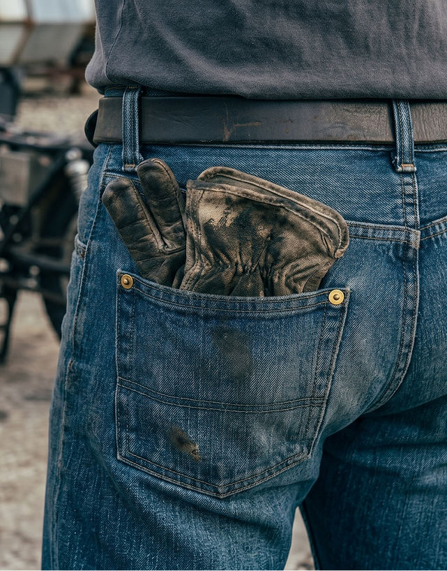 Back pocket of worn blue denim jeans holding a pair of dirty work gloves with a person wearing a brown belt and dark shirt.