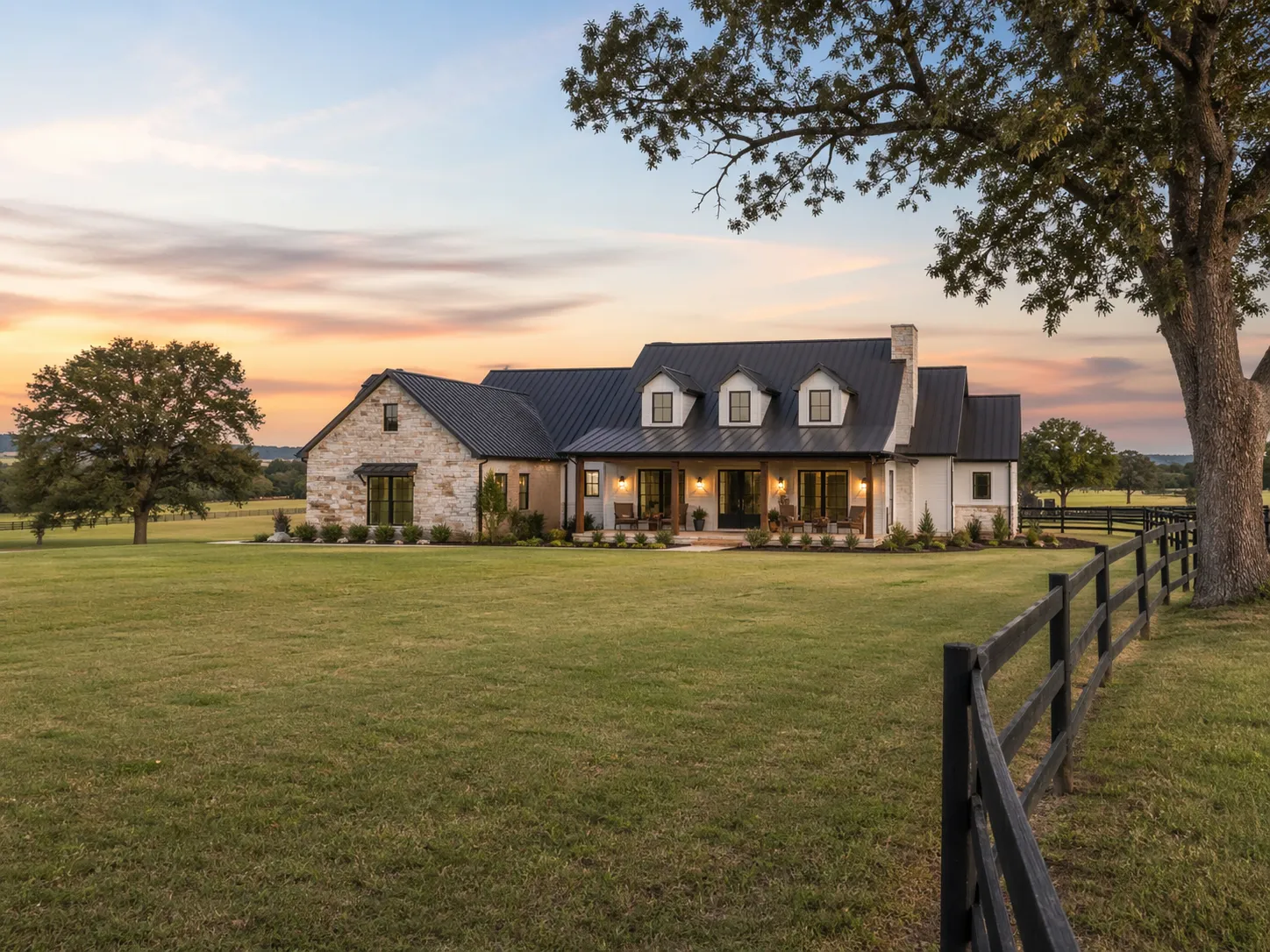 White stone farmhouse with a standing seam metal roof on open acreage at dusk, framed by oaks and a wood fence